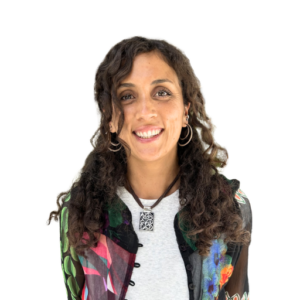 A woman with curly dark hair and a bright smile wears a colorful patterned shirt and a silver pendant necklace.