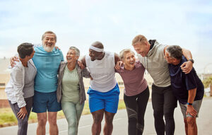 A diverse group of six older adults in athletic wear are standing together with their arms around each other, smiling and laughing.