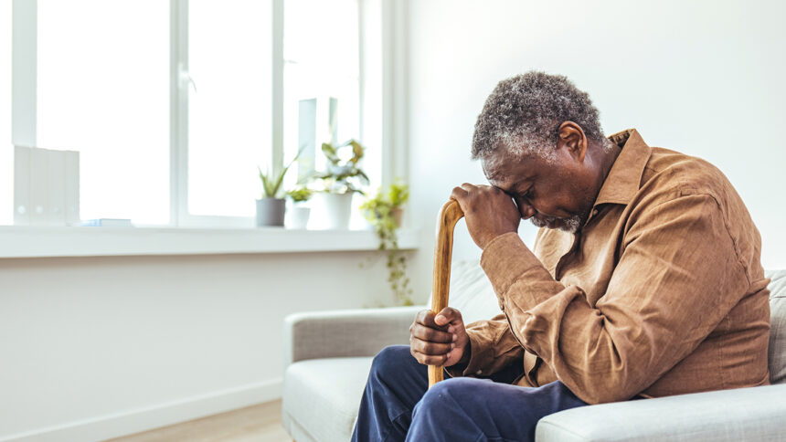 An elderly Black man with graying hair sits on a sofa, head bowed and resting on a cane, appearing distressed.