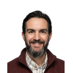 A man with dark hair and a beard smiles at the camera, wearing a maroon quarter-zip pullover over a plaid shirt.
