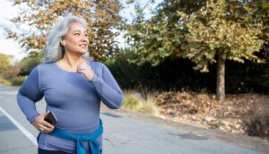 An older woman with gray hair runs on a paved path outdoors, holding a smartphone in her hand.