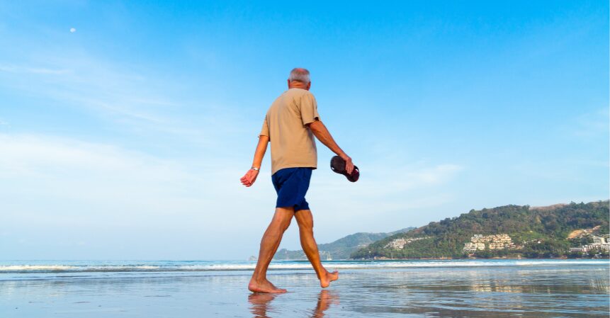 An elderly man walks barefoot on a wet sandy beach holding a flip-flop in his hand.