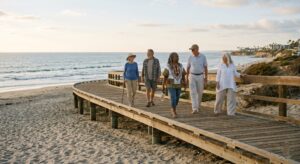 A diverse group of five seniors walks along a wooden boardwalk beside a sandy beach and the ocean.