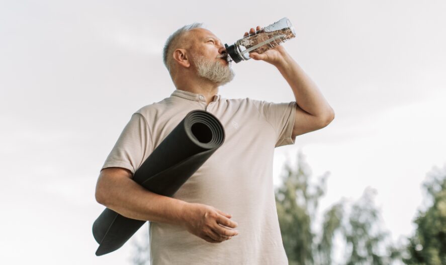 An older man with a grey beard drinks water from a clear bottle while holding a rolled-up yoga mat.