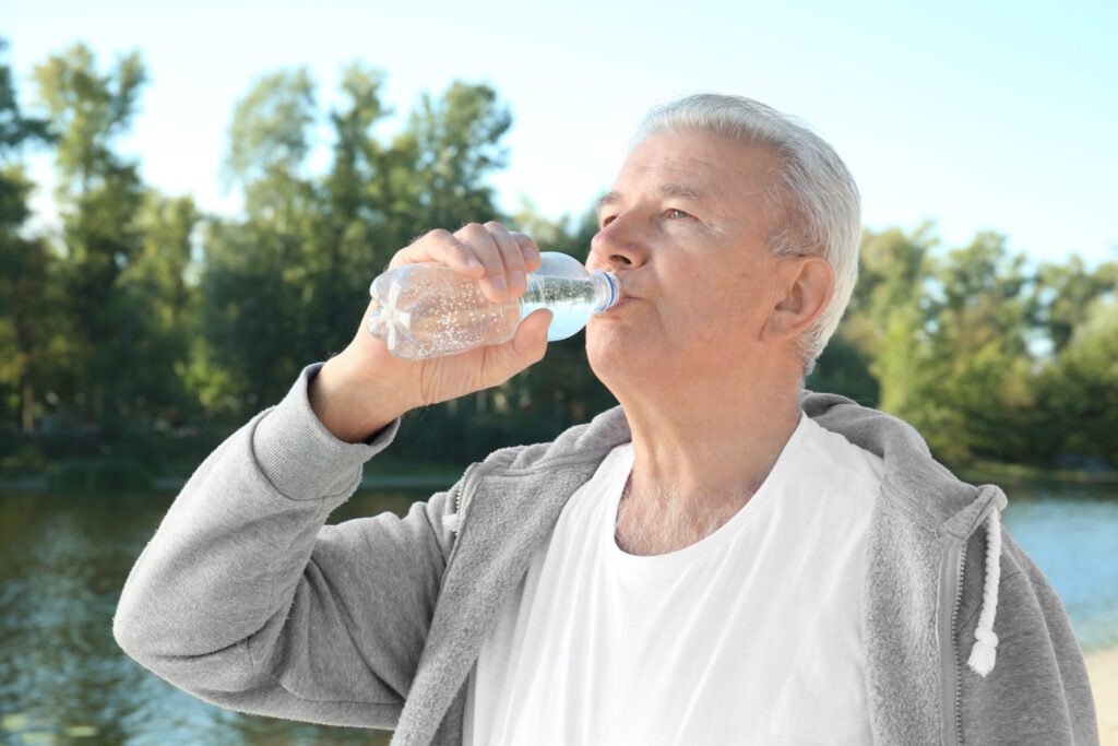 An elderly man with gray hair drinks from a clear plastic bottle of water while wearing a gray zip-up hoodie over a white t-shirt. He is outdoors with trees and a body of water in the background.