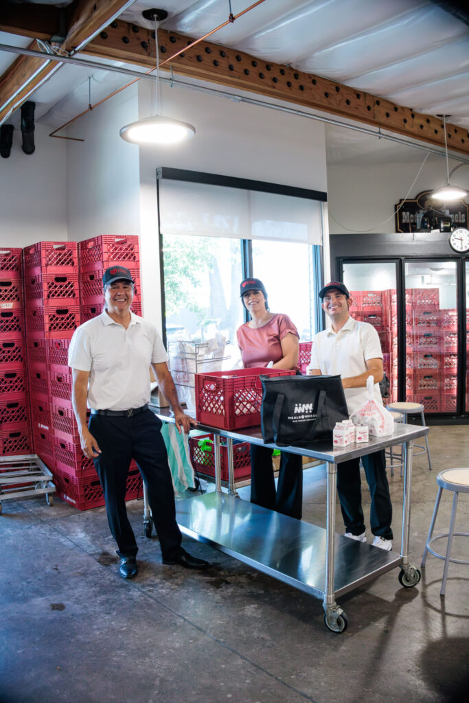 Three people, two men and one woman, stand behind a stainless steel table in a room with red milk crates stacked high. The man on the left wears a white polo shirt, black pants, and a black baseball cap with a red logo. The woman in the middle wears a pinkish-red shirt and a black baseball cap with a red logo. The man on the right wears a white polo shirt, black pants, and a black baseball cap with a red logo. A black tote bag with