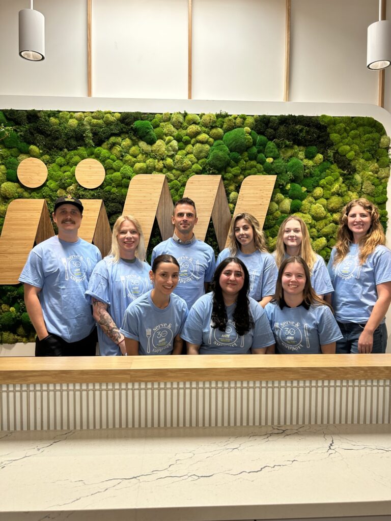 A group of ten people, all wearing light blue t-shirts with the text "serve & support" and a "30" logo, pose for a photo in front of a green moss wall with wooden accents.