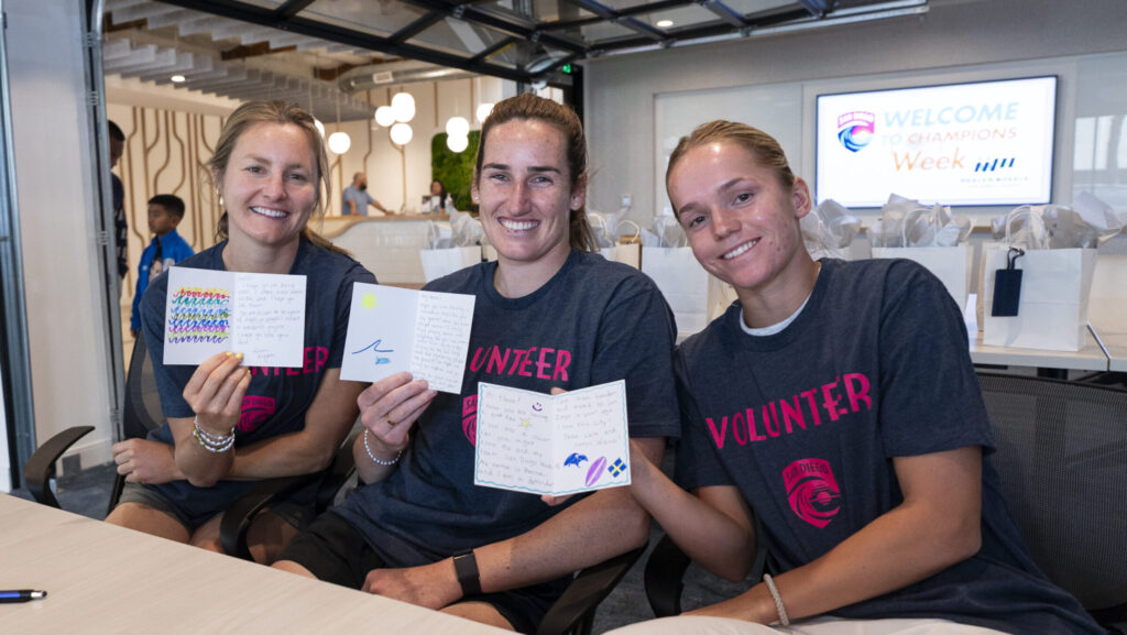 Three women in "Volunteer" t-shirts smile and hold up handmade cards. The woman on the left holds a card with colorful wavy lines. The woman in the middle holds a card with a sun and wave drawing. The woman on the right holds a card with drawings of a wave, a surfboard, and a snowflake. A screen in the background reads "WELCOME TO CHAMPIONS WEEK".