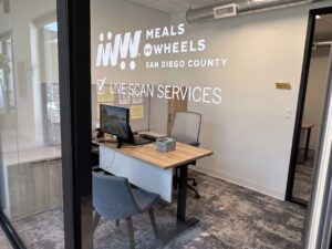 An office reception area with a desk, computer, and chairs, featuring "MEALS ON WHEELS SAN DIEGO COUNTY LIVE SCAN SERVICES" signage on the wall.
