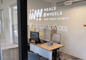 An office reception area with a desk, computer, and chairs, featuring "MEALS ON WHEELS SAN DIEGO COUNTY LIVE SCAN SERVICES" signage on the wall.