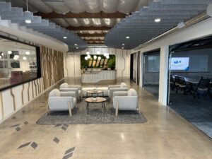 A modern office reception area with a curved reception desk, a green moss wall with a logo, and a seating area with light gray chairs and round coffee tables.