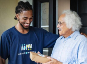A young man in a blue t-shirt with "MEALS ON WHEELS" logo hands a package to an elderly man.